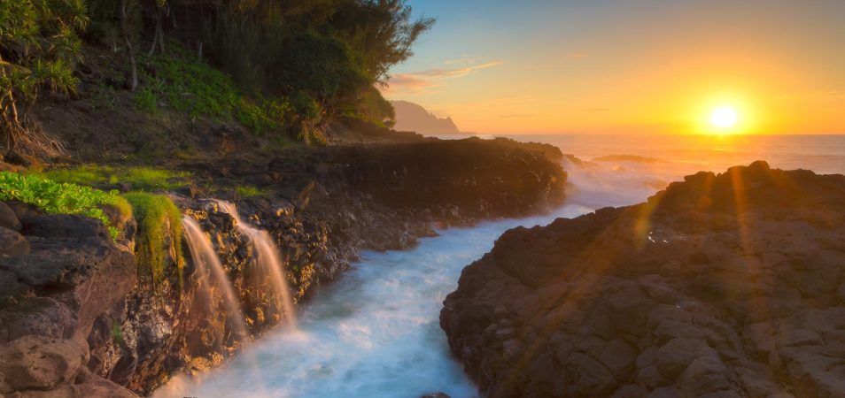 The sun sets over the ocean shining on the rocky coast of Princeville, Kauai in Hawaii