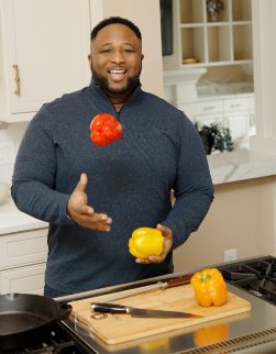 Chef Jernard Wells stands in front of a kitchen stovetop, smiling while holding a yellow bell pepper and throwing a red bell pepper in the air.